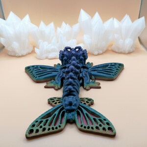 May include: A detailed, blue and green insect sculpture with intricate wing patterns, resting on a light peach surface. Several white, crystal-like formations are in the background. The insect has a textured body and detailed wings.