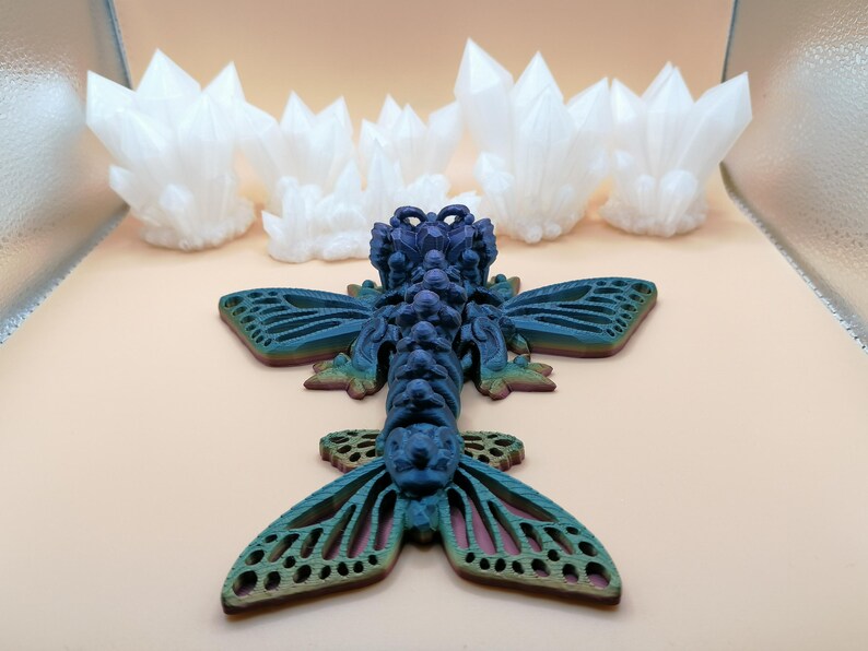 May include: A detailed, blue and green insect sculpture with intricate wing patterns, resting on a light peach surface. Several white, crystal-like formations are in the background. The insect has a textured body and detailed wings.