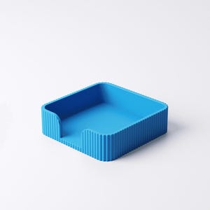 May include: A bright blue, square desk organizer with a ribbed exterior. The organizer has a slot on one side and is designed to hold papers or small items. The solid color and simple design make it a versatile addition to any workspace.