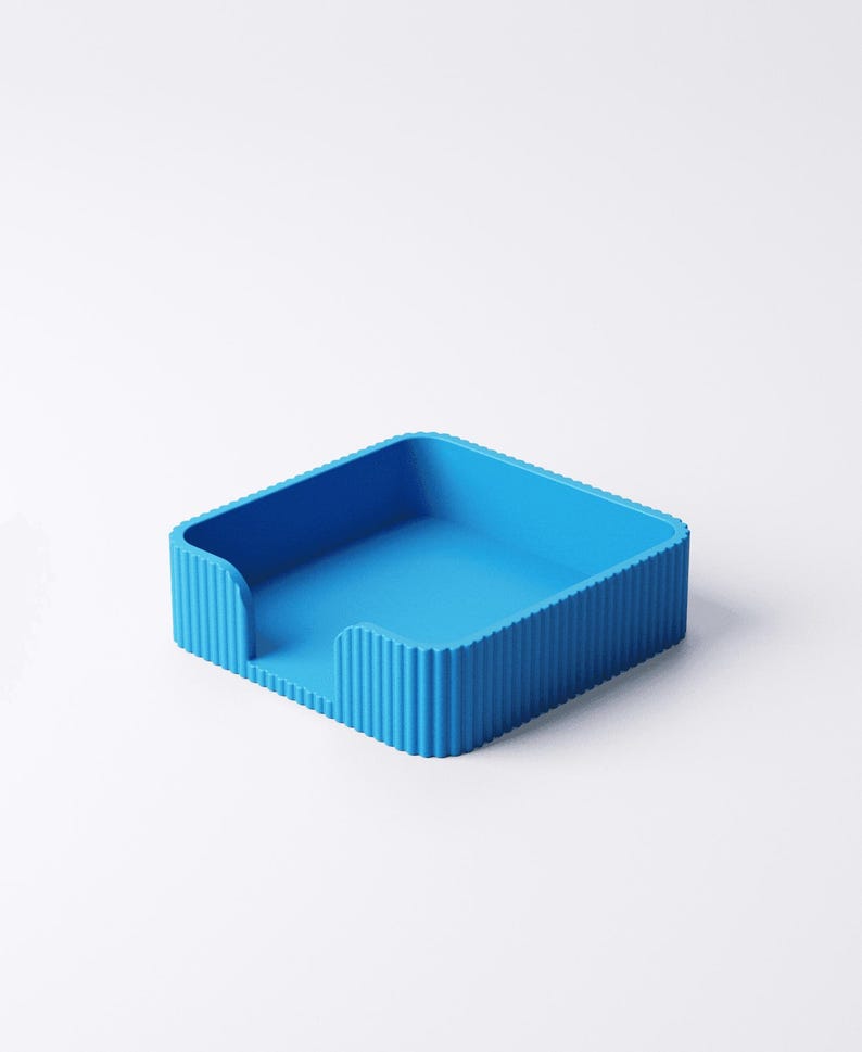 May include: A bright blue, square desk organizer with a ribbed exterior. The organizer has a slot on one side and is designed to hold papers or small items. The solid color and simple design make it a versatile addition to any workspace.