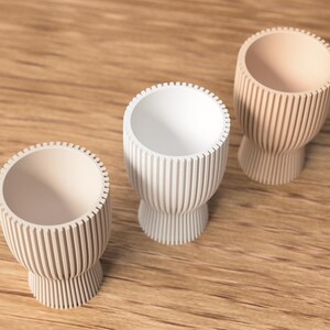 Pu&ograve; includere: Tre piccole portauova decorativi in fila, ognuno con un colore unico. I portauova hanno un design a coste e una base a piedistallo. Uno &egrave; bianco, gli altri due sono beige e marrone chiaro.