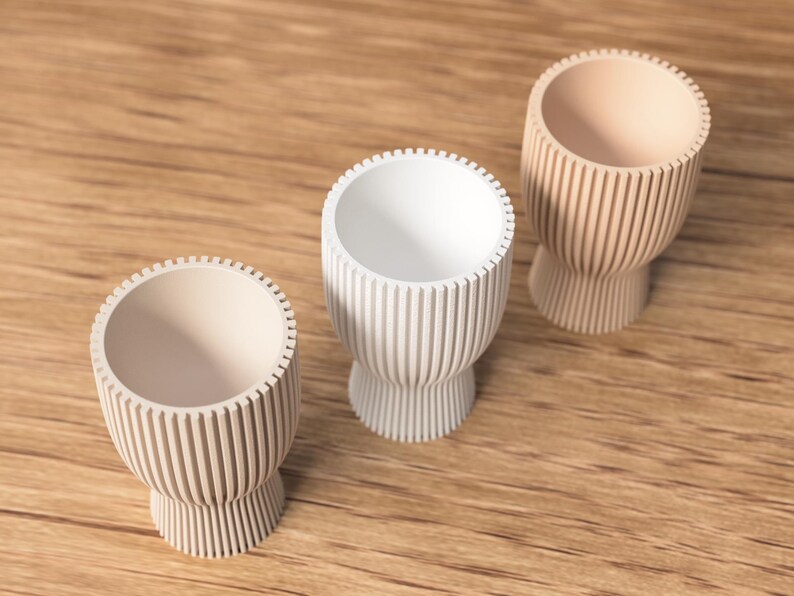 Pu&ograve; includere: Tre piccole portauova decorativi in fila, ognuno con un colore unico. I portauova hanno un design a coste e una base a piedistallo. Uno &egrave; bianco, gli altri due sono beige e marrone chiaro.
