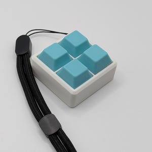 May include: A square, white and light blue keyboard keycap set with four light blue keycaps. The keycaps are arranged in a 2x2 grid. A black lanyard is attached to the keycap set.