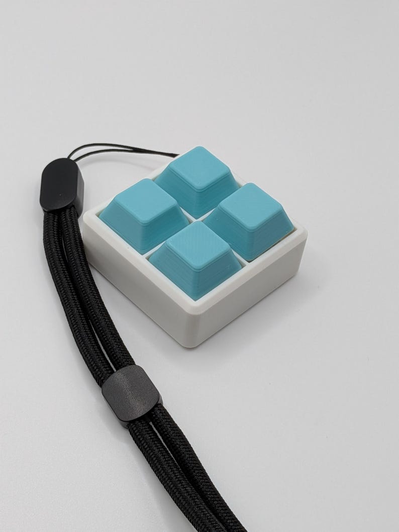 May include: A square, white and light blue keyboard keycap set with four light blue keycaps. The keycaps are arranged in a 2x2 grid. A black lanyard is attached to the keycap set.