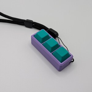 May include: A purple rectangular device with three teal square buttons. The device is attached to a black lanyard. The buttons are slightly raised and have a textured surface. The background is a plain white surface.