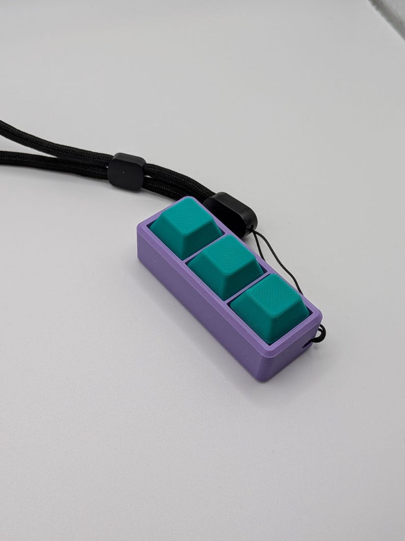 May include: A purple rectangular device with three teal square buttons. The device is attached to a black lanyard. The buttons are slightly raised and have a textured surface. The background is a plain white surface.