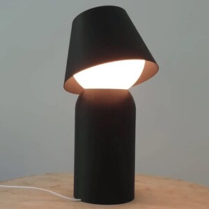 May include: A modern black table lamp with a unique design. The lamp features a cylindrical base and a tilted lampshade, revealing a bright, glowing bulb. The lamp is on a wooden surface.
