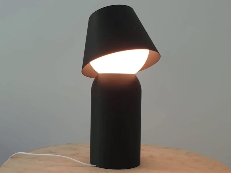 May include: A modern black table lamp with a unique design. The lamp features a cylindrical base and a tilted lampshade, revealing a bright, glowing bulb. The lamp is on a wooden surface.