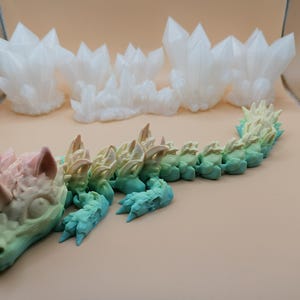 Puede incluir: Un juguete de drag&oacute;n articulado en colores pastel, con un degradado de rosa a azul. El drag&oacute;n est&aacute; tumbado, con una cabeza detallada y una espalda puntiaguda. En el fondo hay varias formaciones de cristales blancos.