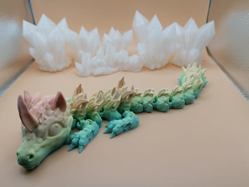 Puede incluir: Un juguete de drag&oacute;n articulado en colores pastel, con un degradado de rosa a azul. El drag&oacute;n est&aacute; tumbado, con una cabeza detallada y una espalda puntiaguda. En el fondo hay varias formaciones de cristales blancos.