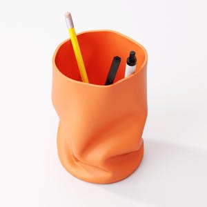 May include: Orange desk organizer with a unique, crumpled design. A yellow pencil, a black pen, and a white marker are inside. The container is made of a solid, opaque material and sits on a white surface.