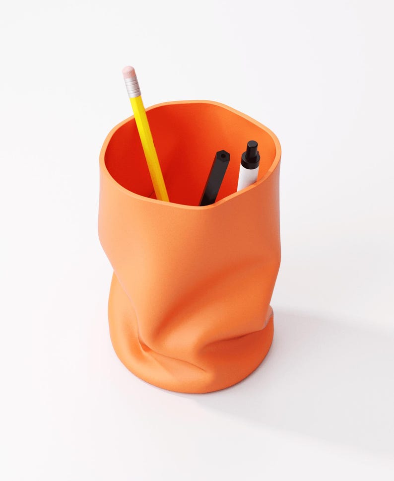 May include: Orange desk organizer with a unique, crumpled design. A yellow pencil, a black pen, and a white marker are inside. The container is made of a solid, opaque material and sits on a white surface.