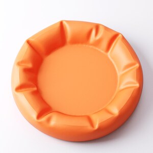 May include: An orange, round ashtray with a smooth, matte finish. The ashtray has a central recessed area and a series of raised, curved edges around the perimeter. The color is a vibrant, solid orange.