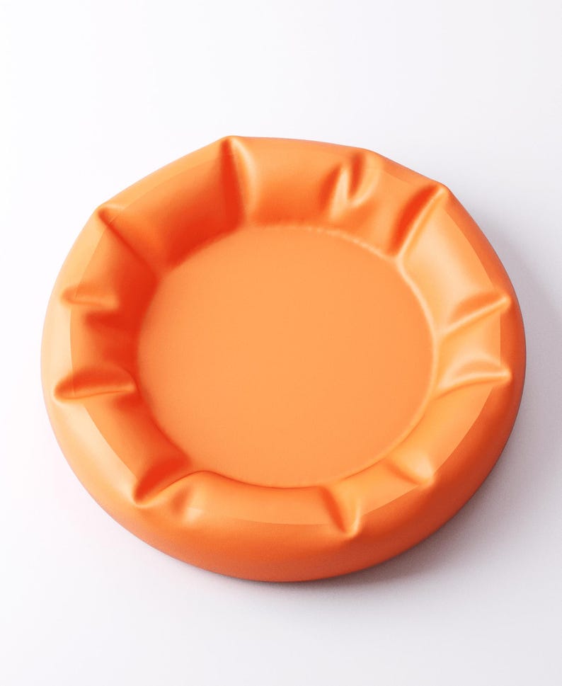 May include: An orange, round ashtray with a smooth, matte finish. The ashtray has a central recessed area and a series of raised, curved edges around the perimeter. The color is a vibrant, solid orange.