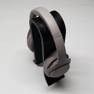 K&ouml;nnte beinhalten: Graue Over-Ear-Kopfh&ouml;rer auf einem schwarzen Kopfh&ouml;rerst&auml;nder. Die Kopfh&ouml;rer haben ein gepolstertes Kopfband und Ohrmuscheln. Der St&auml;nder ist ein geschwungenes, minimalistisches Design. Der Hintergrund ist schlicht wei&szlig;.
