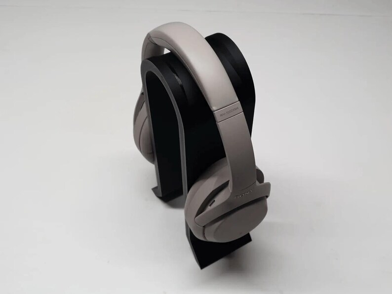 K&ouml;nnte beinhalten: Graue Over-Ear-Kopfh&ouml;rer auf einem schwarzen Kopfh&ouml;rerst&auml;nder. Die Kopfh&ouml;rer haben ein gepolstertes Kopfband und Ohrmuscheln. Der St&auml;nder ist ein geschwungenes, minimalistisches Design. Der Hintergrund ist schlicht wei&szlig;.