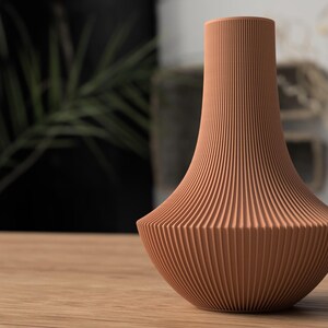 May include: A terracotta-colored vase with a ribbed, textured surface. The vase has a wide base that tapers to a narrow neck. The vase is sitting on a wooden surface.