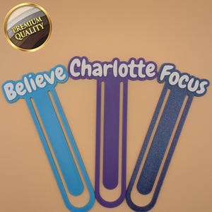 May include: Three flat, elongated paperclip-shaped bookmarks with rounded ends. The left one is light blue with the word Believe, the centre is purple with the name Charlotte, and the right one is dark blue glitter with the word Focus.