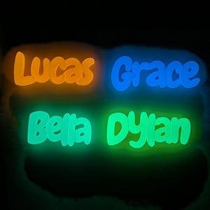 May include: Four illuminated name signs: Lucas in orange, Grace in blue, Bella in green, and Dylan in green. The names are in a playful font, set against a dark background.
