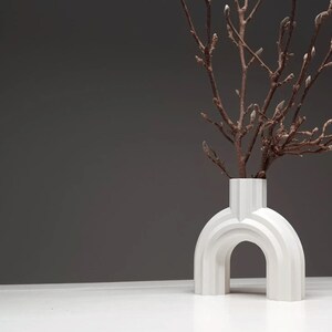 May include: A modern white vase with an arched design, holding branches with budding flowers. The vase has a unique architectural shape and sits on a white surface against a gray backdrop. A decorative home accent.