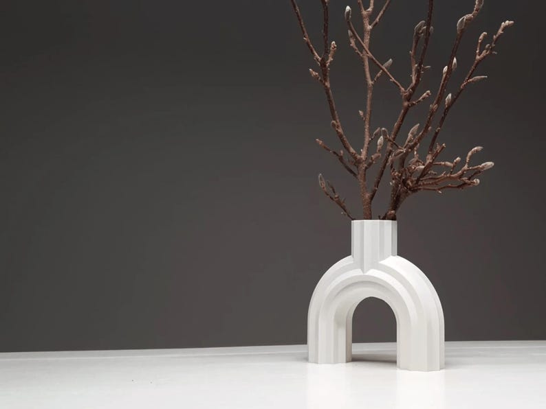 May include: A modern white vase with an arched design, holding branches with budding flowers. The vase has a unique architectural shape and sits on a white surface against a gray backdrop. A decorative home accent.