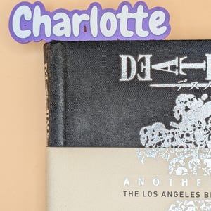 May include: A purple and white 3D printed bookmark featuring the name Charlotte in a rounded font, positioned at the top edge of a black book with silver text that reads Death Note and a beige paper band with the text Another The Los Angeles Bi.