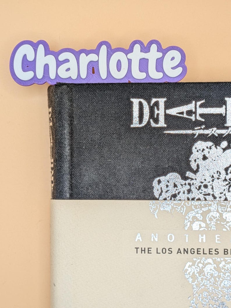 May include: A purple and white 3D printed bookmark featuring the name Charlotte in a rounded font, positioned at the top edge of a black book with silver text that reads Death Note and a beige paper band with the text Another The Los Angeles Bi.
