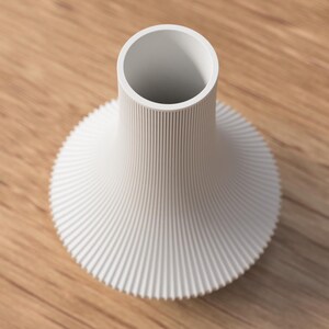 May include: A white, modern vase with a cylindrical neck and a flared, ridged base. The vase has a minimalist design and is set against a wooden surface.