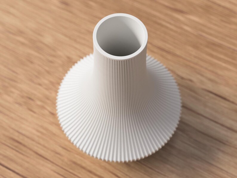 May include: A white, modern vase with a cylindrical neck and a flared, ridged base. The vase has a minimalist design and is set against a wooden surface.