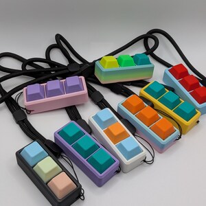 May include: A collection of colorful keycap pendants, each featuring three keycaps in a row. The keycaps are in various colors, including pink, purple, blue, green, yellow, orange, and red. Each pendant has a black lanyard.