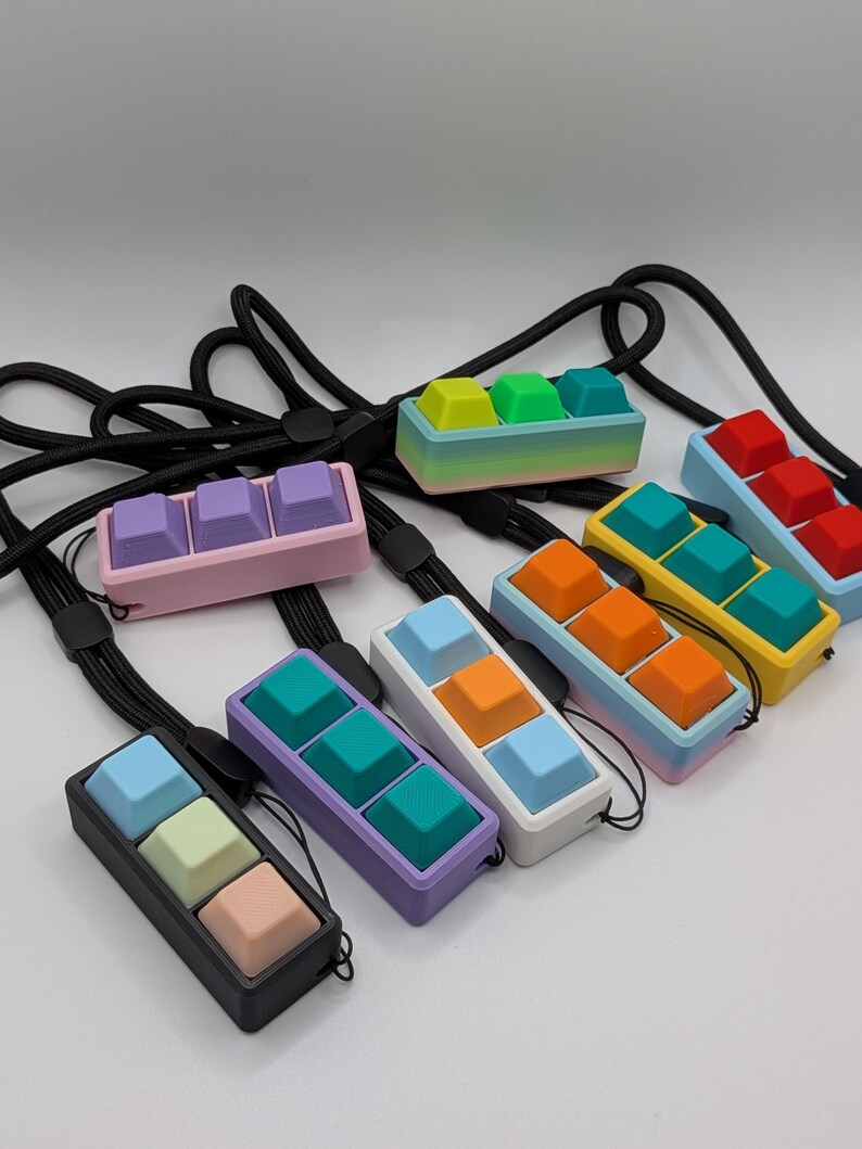 May include: A collection of colorful keycap pendants, each featuring three keycaps in a row. The keycaps are in various colors, including pink, purple, blue, green, yellow, orange, and red. Each pendant has a black lanyard.