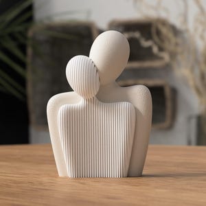 Op de afbeelding: Een cr&egrave;mekleurige sculptuur die twee abstracte figuren omarmend afbeeldt. De grotere figuur heeft een glad oppervlak, terwijl de kleinere figuur en de voorkant van de grotere figuur verticale lijnen hebben. De sculptuur staat op een houten oppervlak.