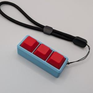 May include: A custom mechanical keyboard macro pad with three red keycaps set in a light blue frame. A black lanyard is attached for portability. The keycaps are square and textured.