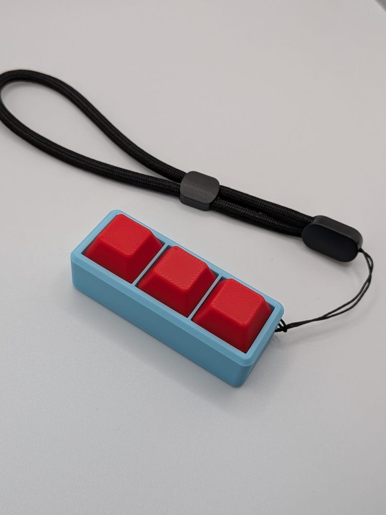 May include: A custom mechanical keyboard macro pad with three red keycaps set in a light blue frame. A black lanyard is attached for portability. The keycaps are square and textured.
