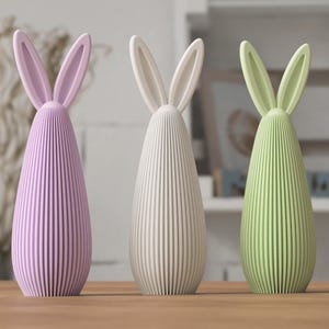 May include: Three decorative bunny figurines in pastel colors. The figurines have a ribbed, teardrop-shaped body and long, upright ears. The colors are lavender, off-white, and light green. They are displayed on a wooden surface.