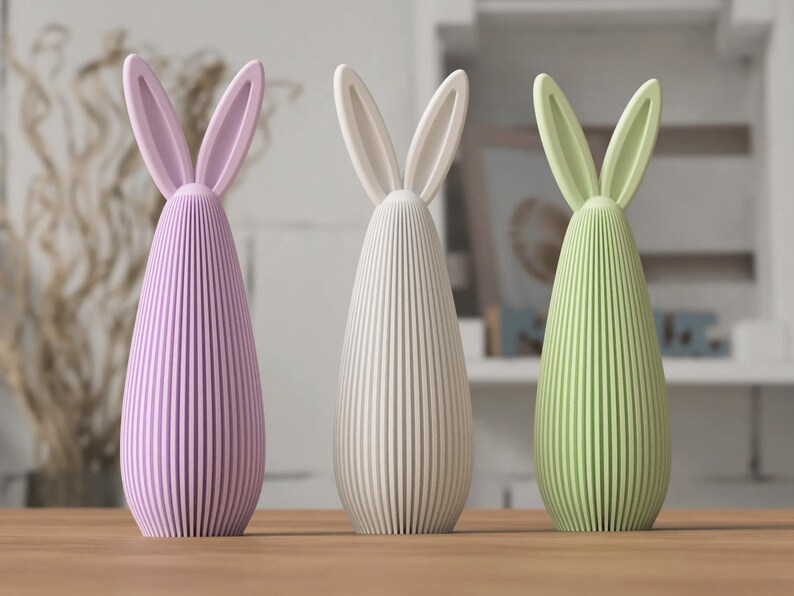 May include: Three decorative bunny figurines in pastel colors. The figurines have a ribbed, teardrop-shaped body and long, upright ears. The colors are lavender, off-white, and light green. They are displayed on a wooden surface.
