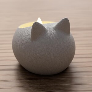 May include: A gray, cat-shaped candle holder with a textured surface. The candle holder has two pointed ears and a round body. The inside of the holder is yellow, and a white candle is lit inside.