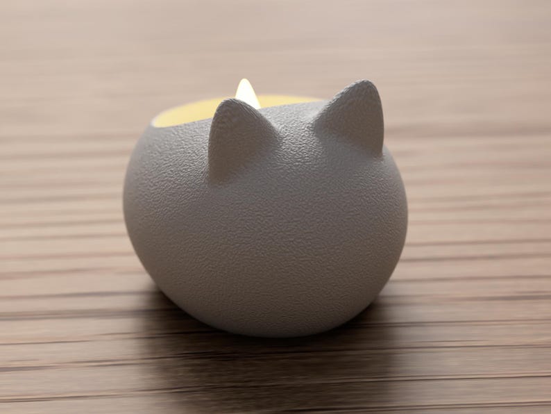 May include: A gray, cat-shaped candle holder with a textured surface. The candle holder has two pointed ears and a round body. The inside of the holder is yellow, and a white candle is lit inside.