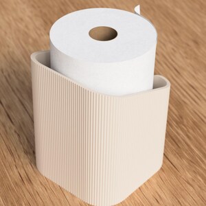 Op de afbeelding: Een beige toiletpapierhouder met een verticaal geribbeld ontwerp, met een rol wit toiletpapier. De houder heeft een gebogen bovenkant en een vierkante basis. De achtergrond is een houten oppervlak.