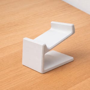May include: A white, minimalist phone stand with a slanted top and a rectangular base. The stand is made of a solid material and sits on a light brown wooden surface.
