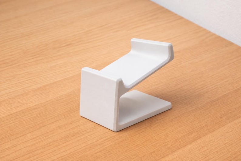 May include: A white, minimalist phone stand with a slanted top and a rectangular base. The stand is made of a solid material and sits on a light brown wooden surface.