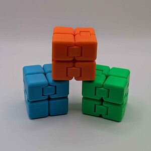 May include: Three colorful infinity cubes in orange, blue, and green are arranged on a white surface. Each cube is composed of smaller, connected blocks that can be flipped and rotated. The cubes are designed for fidgeting and stress relief.