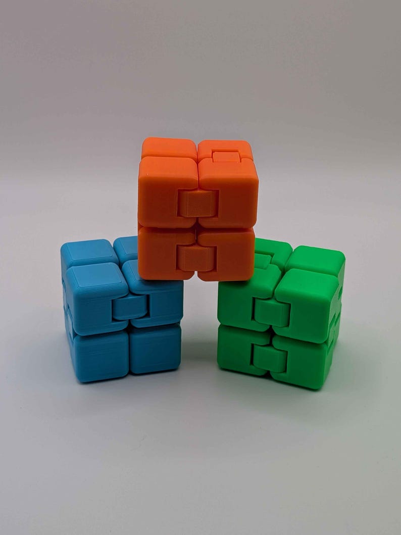 May include: Three colorful infinity cubes in orange, blue, and green are arranged on a white surface. Each cube is composed of smaller, connected blocks that can be flipped and rotated. The cubes are designed for fidgeting and stress relief.