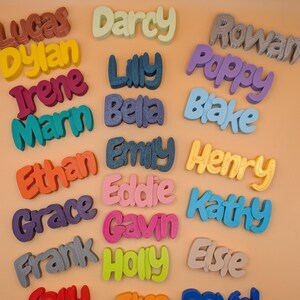 May include: A selection of brightly coloured 3D-printed name signs. Names like Lucas, Dylan, and Irene are displayed in a playful font. The signs are in various colours, including red, yellow, blue, and green, and are arranged on a light orange surface.
