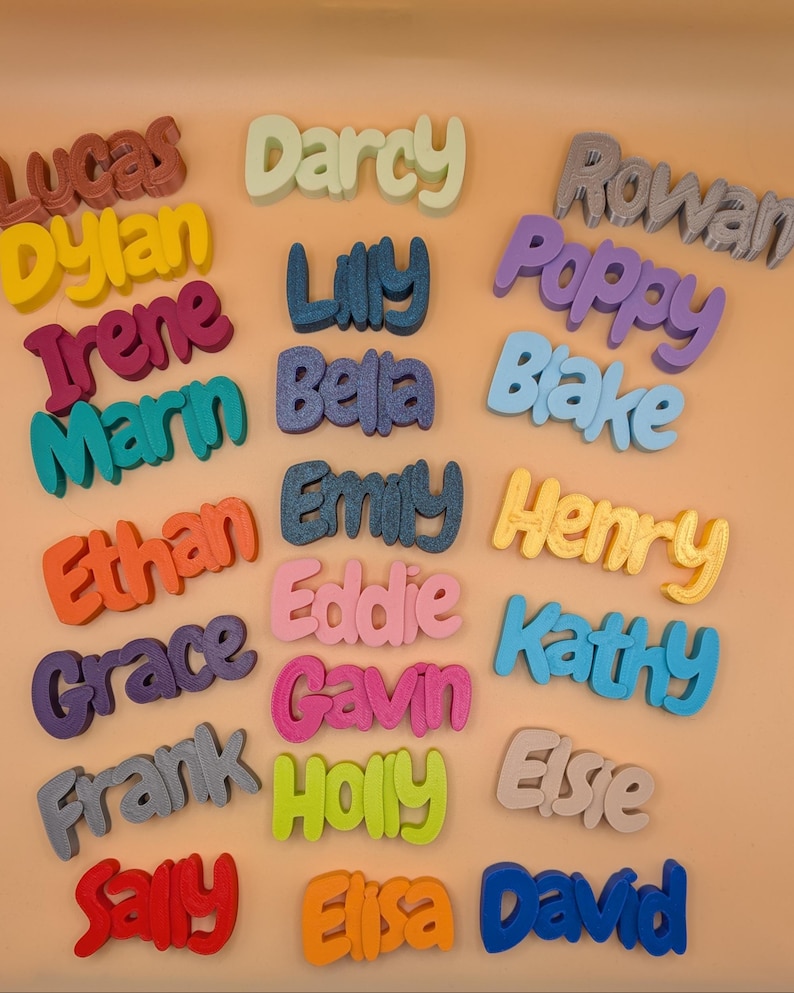 May include: A selection of brightly coloured 3D-printed name signs. Names like Lucas, Dylan, and Irene are displayed in a playful font. The signs are in various colours, including red, yellow, blue, and green, and are arranged on a light orange surface.