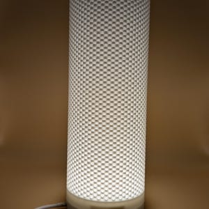 K&ouml;nnte beinhalten: Zylindrische Lampe mit einem strukturierten, karierten Muster. Die Lampe ist wei&szlig; und strahlt ein weiches, warmes Licht aus. Der Sockel ist ein hellerer Wei&szlig;ton, und ein wei&szlig;es Kabel erstreckt sich von unten.