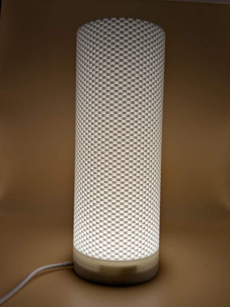K&ouml;nnte beinhalten: Zylindrische Lampe mit einem strukturierten, karierten Muster. Die Lampe ist wei&szlig; und strahlt ein weiches, warmes Licht aus. Der Sockel ist ein hellerer Wei&szlig;ton, und ein wei&szlig;es Kabel erstreckt sich von unten.