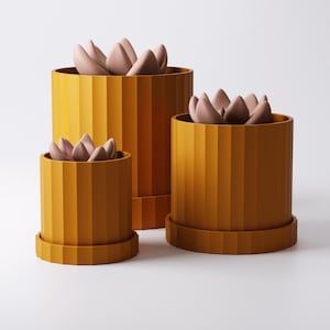 Puede incluir: Tres macetas cil&iacute;ndricas de color amarillo mostaza con texturas acanaladas verticales, cada una con una base circular a juego. Dentro de cada maceta hay una planta suculenta estilizada con p&eacute;talos de color rosa p&aacute;lido, lisos y redondeados.
