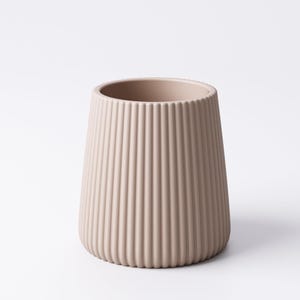 May include: A beige, ribbed ceramic vase with a wide opening and a tapered shape. The vertical ridges create a textured surface. The vase is a neutral color and stands on a white background.