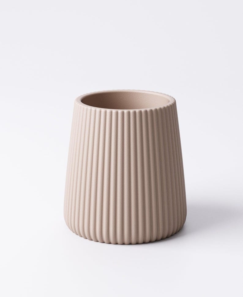 May include: A beige, ribbed ceramic vase with a wide opening and a tapered shape. The vertical ridges create a textured surface. The vase is a neutral color and stands on a white background.
